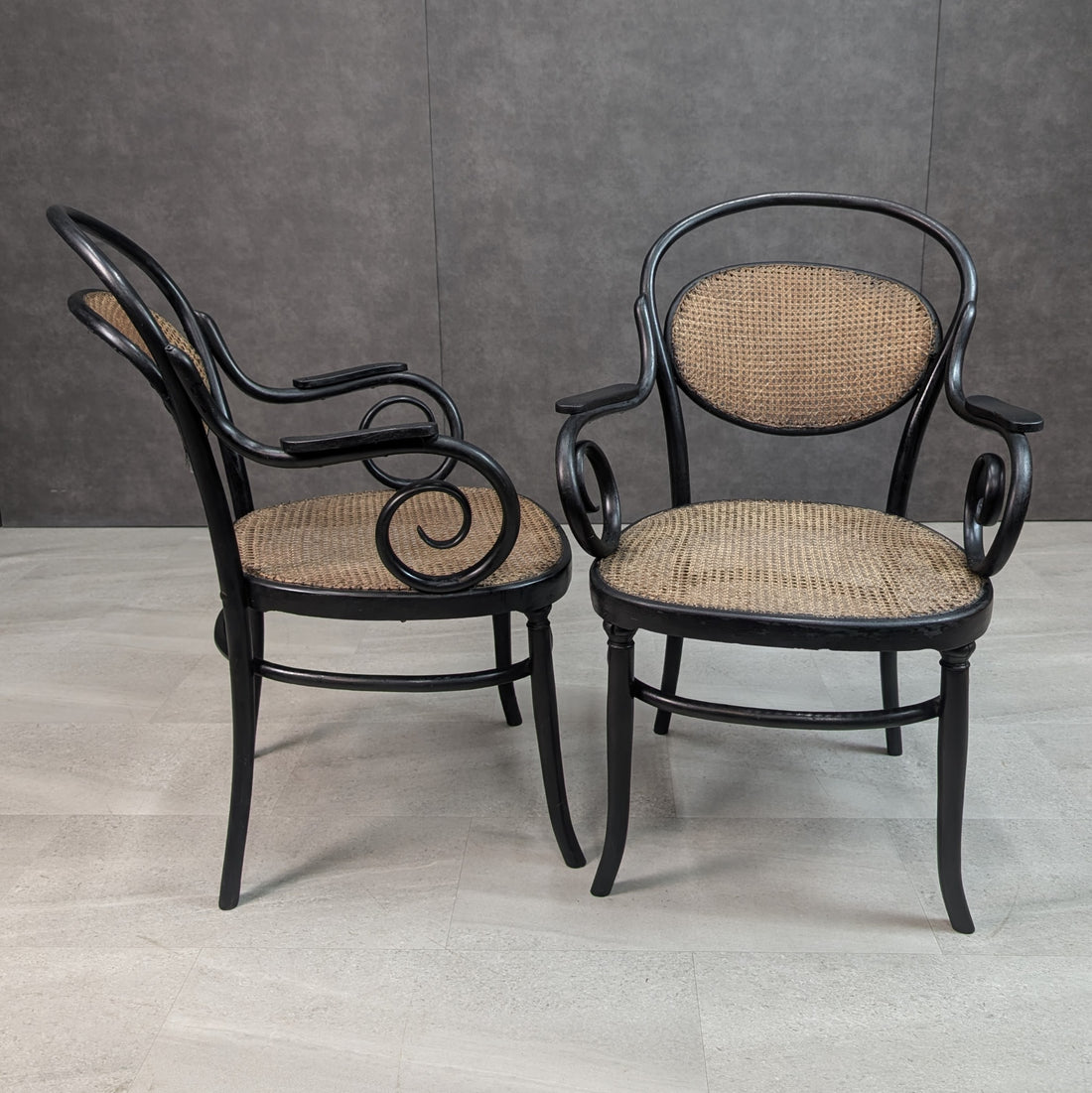 Three quarter view of two antique black bentwood and cane armchairs showing scroll arms and oval backs